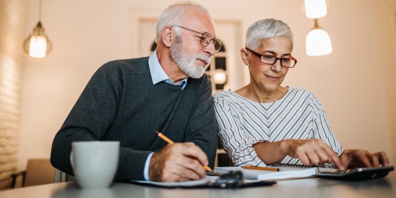 Two people sit at a table in a well-lit room. One writes with a pencil, the other uses a laptop. Hanging lights are visible above, suggesting a collaborative work setting.
