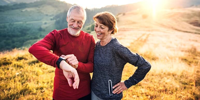 An elderly couple out on a hike, with the husband lifting his wrist as both of them check out the recorded data on his insured fitness watch. 