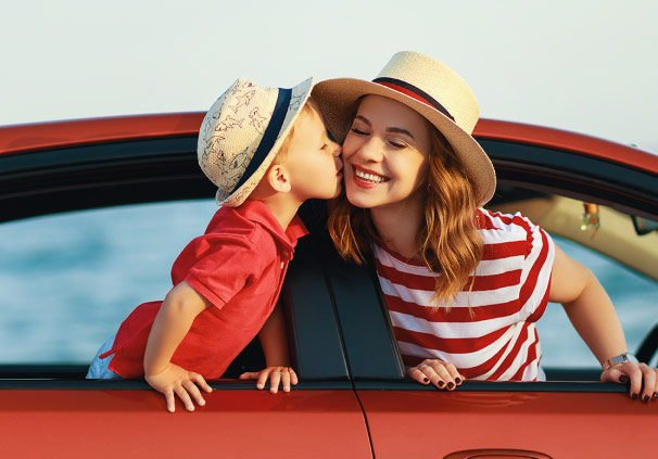 A young toddler sits in the back of the car while his mom sits in the front. Both are leaning out the window, and the toddler is kissing his mom on the cheek.