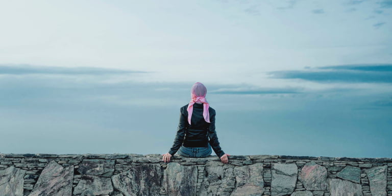 A lady wearing a black leather jacket and blue jeans and a pink bandana wrapped around her head sitting on a stone wall looking at the cloudy horizon in front of her.