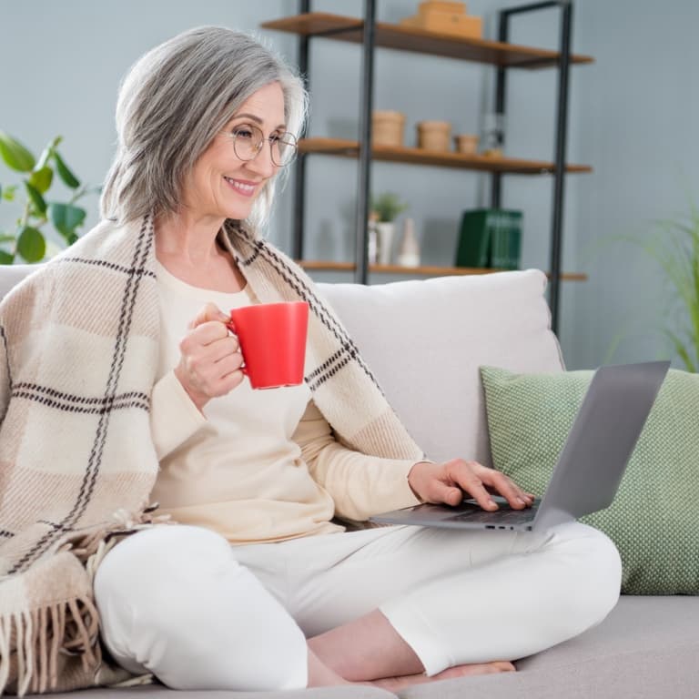 A mature woman relaxing on the couch, enjoying a cup of coffee while looking at her laptop.