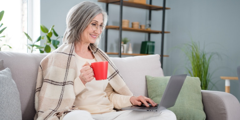 A woman with long grey hair holding a red mug and working on her laptop on the couch.