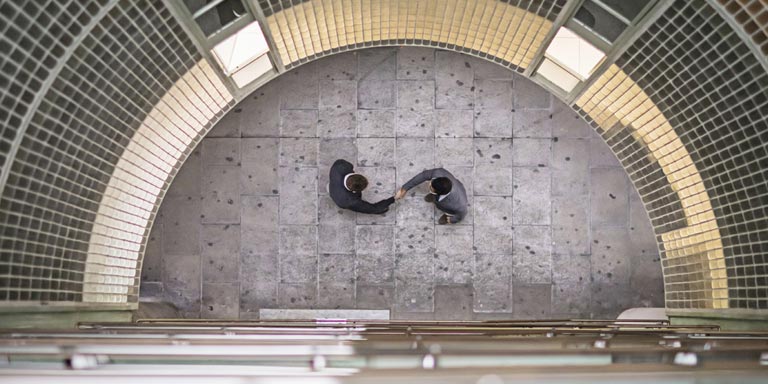 An aerial view of the inside of a round building, with two businessmen on the ground floor, shaking hands.