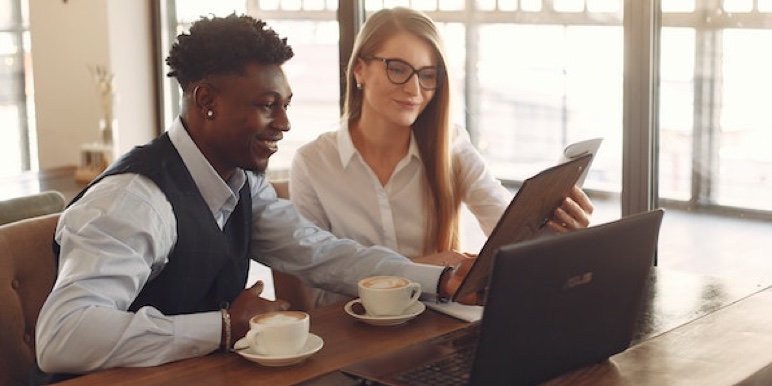 A young African man and woman sit at a table with 2 cappucinos and an open laptop.