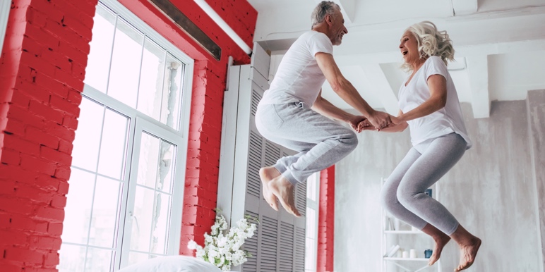 An older couple wearing white t-shirts and grey pants holding hands and jumping on their bed excitedly.