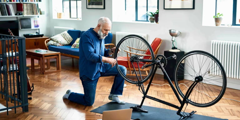 Old man in his blue overalls fixing his bicycle that's placed upside down in his living room.