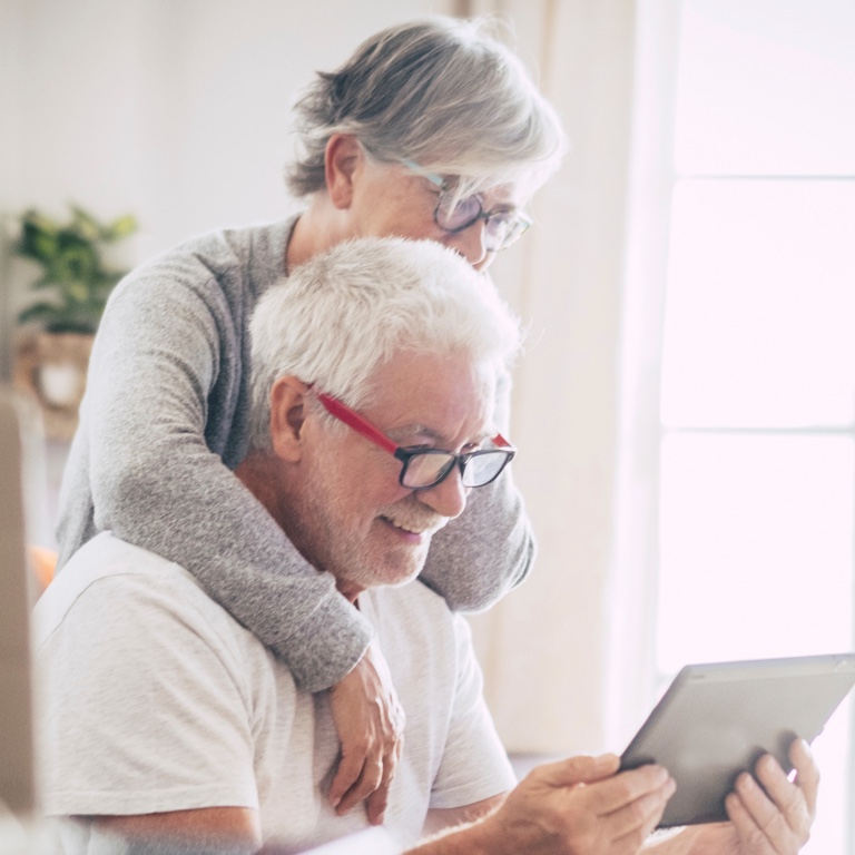 An elderly couple looking into a tablet. The wife is standing over the husband’s shoulder. 
