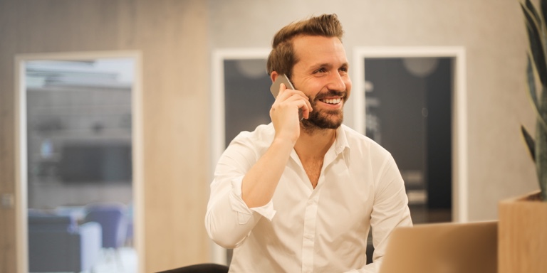 A young man in a white shirt with his sleeves rolled up to the middle of his forearm, speaking on his cell phone and smiling.