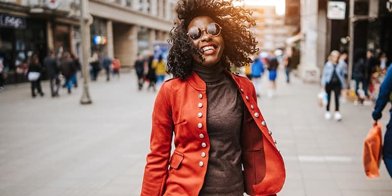 A young woman with curly hair and wearing a red jacket and sunglasses walking in a street.