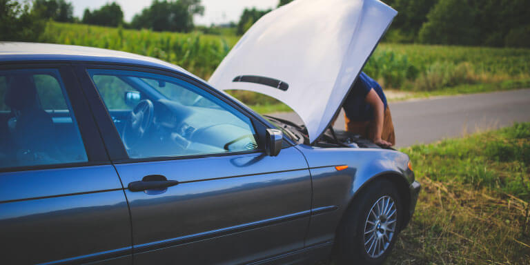 Stranded driver inspecting a car with an open hood on the side of a road. The breakdown highlights the need for roadside assistance and emergency support from business insurance.
