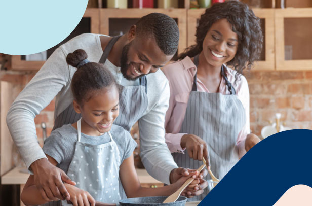 A smiling African man in an apron at home in the kitchen showing his daughter how to prepare food, while helping her mother cook. 