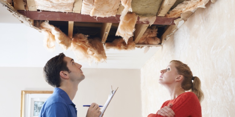 A lady in a red jersey stands opposite her claims assessor, holding a clipboard, looking at her ceiling damage after a heavy storm. The lady has home insurance with Momentum Insure.