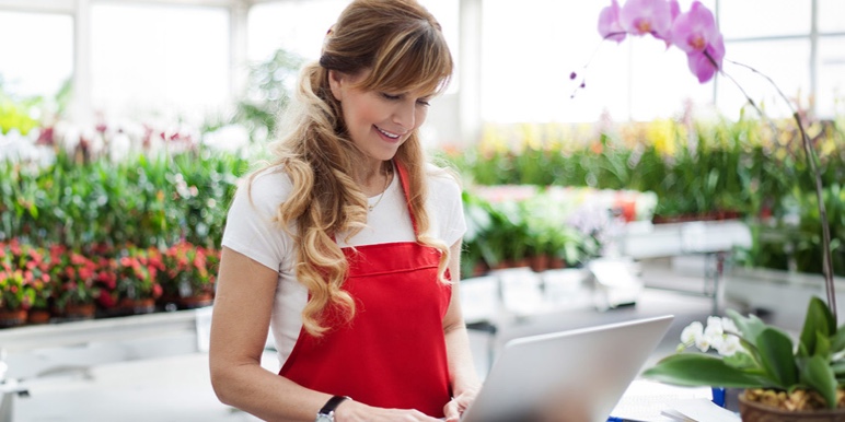 A lady standing in a nursery with flowers in the background working on a laptop, wearing a red apron.