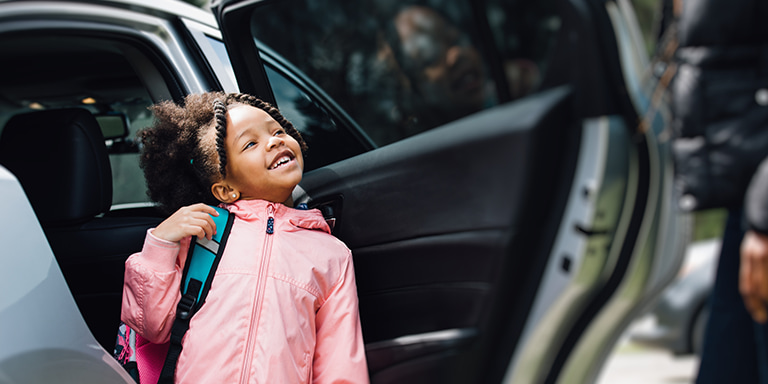 A happy little girl in a pink rain jacket, carrying a school bag,  standing outside of an open car door, confident that she is  safe and secure.