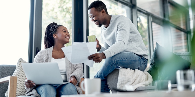 A happy, young couple, sitting on the couch while discussing their retirement fund beneficiaries.
