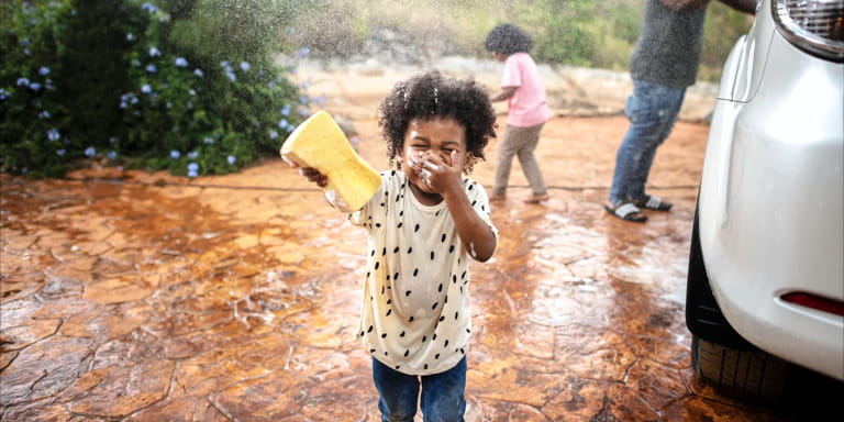 A dad is washing his car with his two young daughters while splashing water on each other.