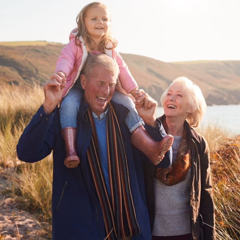 An older couple taking a walk in nature. The grandfather is holding his granddaughter on his shoulders.