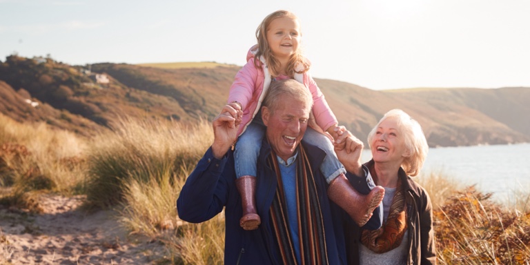An older couple taking a walk in nature. The grandfather is holding his granddaughter on his shoulders.