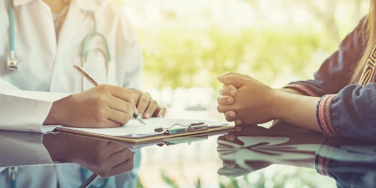 A doctor and patient sit across a desk in consultation while the doctor writes down notes.