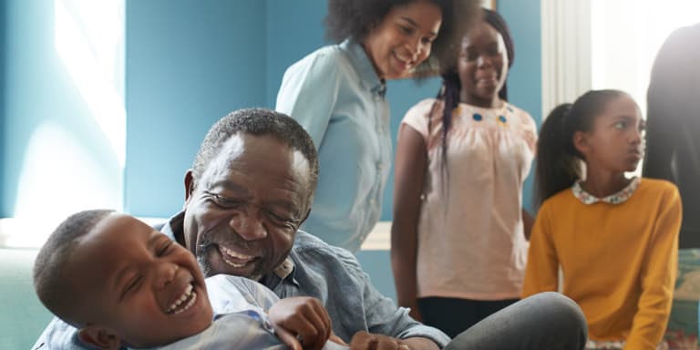 A grandfather tickles and embraces his grandson while his daughter and granddaughter look at them smiling.