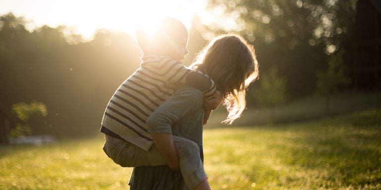 Happy mom holds her toddler son on her back against a background of trees in summer with the late afternoon sun shining through the trees.