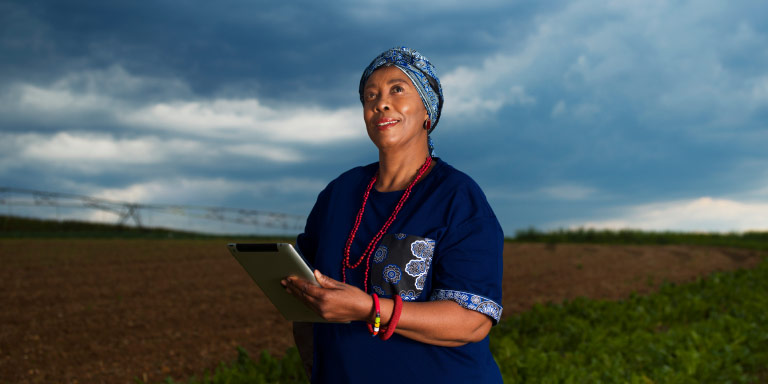 A middle aged woman dressed in African attire and holding a tablet, outside an open field, looking onwards.