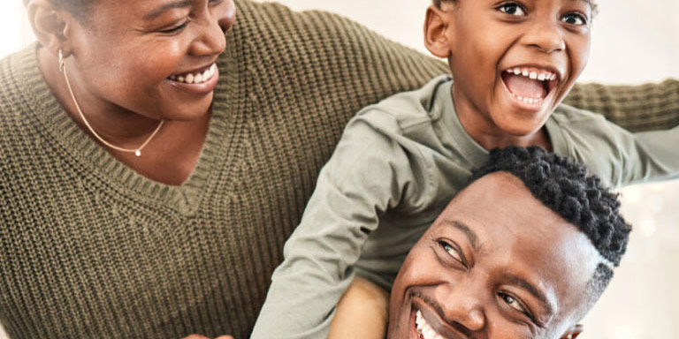 Black family of three with the little boy on his father’s shoulders and the mother looking at them smiling.