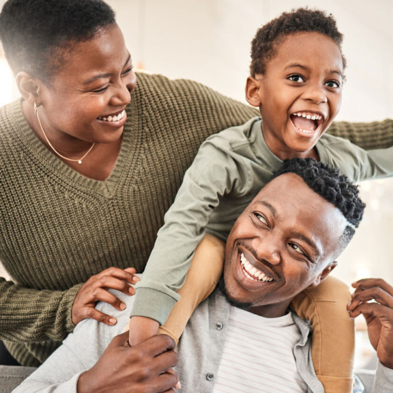 Black family of three with the little boy on his father’s shoulders and the mother looking at them smiling.