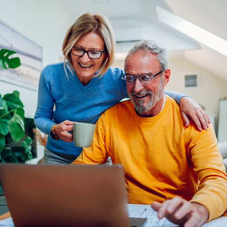 A smiling middle-aged woman standing over her husband’s shoulder, with coffee in hand, while they review the terms and eligibility of his retirement annuity on the computer screen for two-pot.