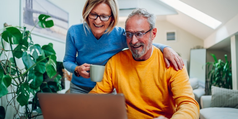 A smiling middle-aged woman standing over her husband’s shoulder, with coffee in hand, while they review the terms and eligibility of his retirement annuity on the computer screen for two-pot.