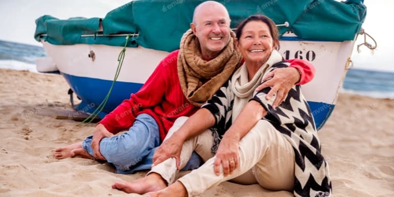 A retired couple sits on the sandy beach, with the man’s arm wrapped around her. She’s wearing a cozy poncho, and behind them, is a sailboat.