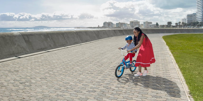 A mum teaches her son to ride a bike on the pavement outside the coastal big city on a slightly overcast day.