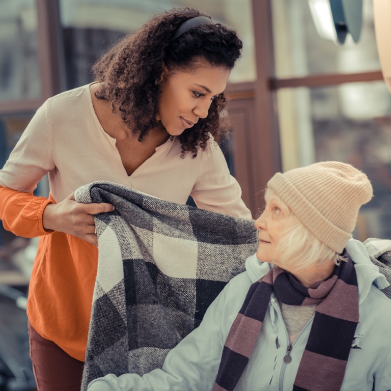 Brown woman putting a checkered blanket around the shoulders of an old white woman. 