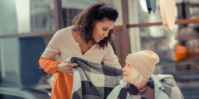 Brown woman putting a checkered blanket around the shoulders of an old white woman. 