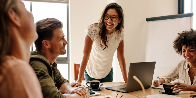 Four colleagues with black coffee mugs laughing around a table at a work meeting.