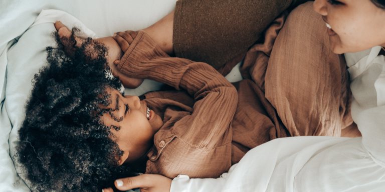 A young woman cheerfully waking up her daughter for the day, who is still in bed.