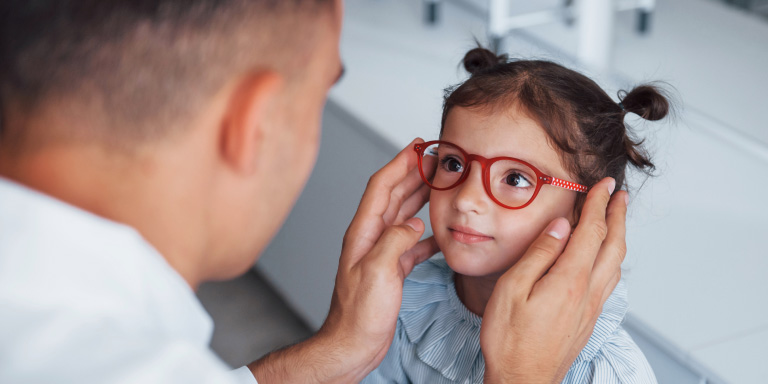A cute young girl smiles while an optometrist holds out red-framed glasses for her to try on.