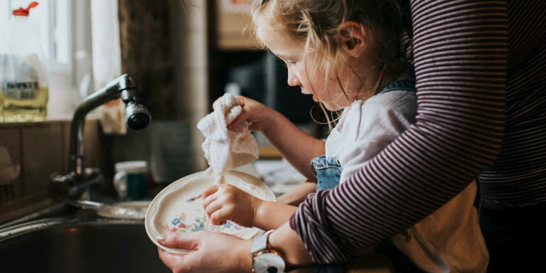 A little girl helps her mum dry a pretty patterned side plate after it being washed at the sink in the kitchen.