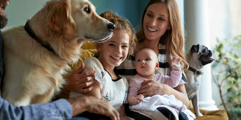 A mum sits with her two daughters on a couch with their golden retriever and fawn pug.