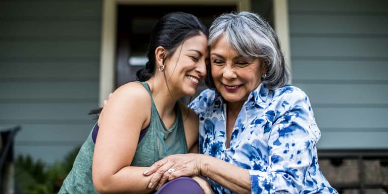 A mum and daughter embrace each other while sitting on the steps on the porch in front of their home.