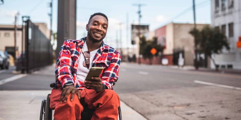 A young man in orange trousers and red checkered jacket sitting in a wheelchair in an empty street, smiling.