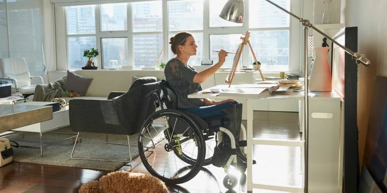 A young lady in a wheelchair sitting at her drawing desk, painting on an easel. She is in her modern apartment with lots of natural light and her dog is lying on the floor beside her.