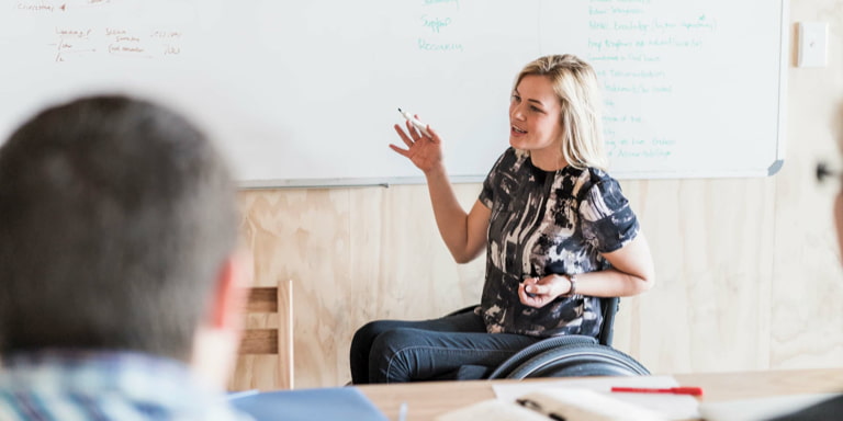 A young female teacher in a wheelchair using a white board marker to expalin a concept to her students in her class.