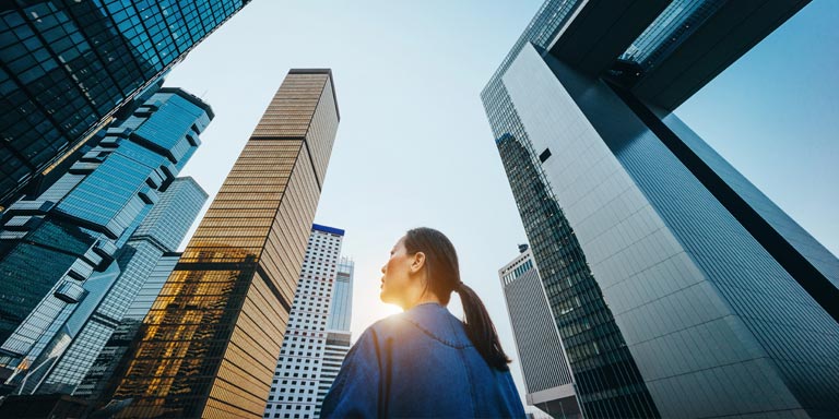 A woman in a blue top looking up at numerous modern, glass skyscrapers.