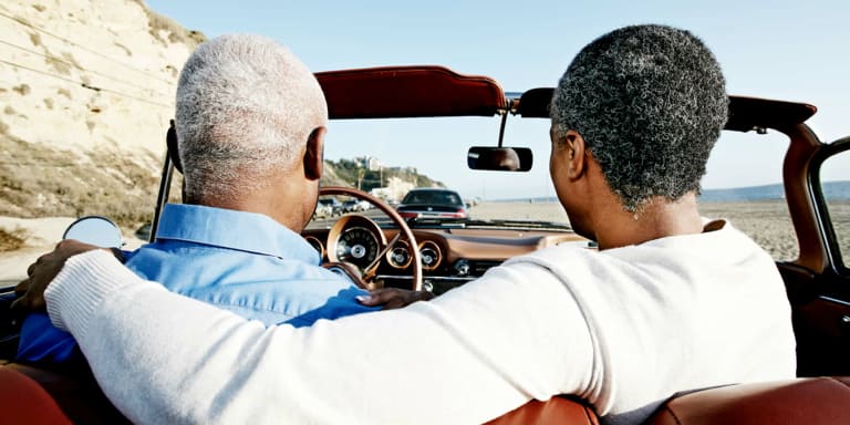An old couple taking a ride in a vintage drop top car. The woman has her arm around her husband who's driving.