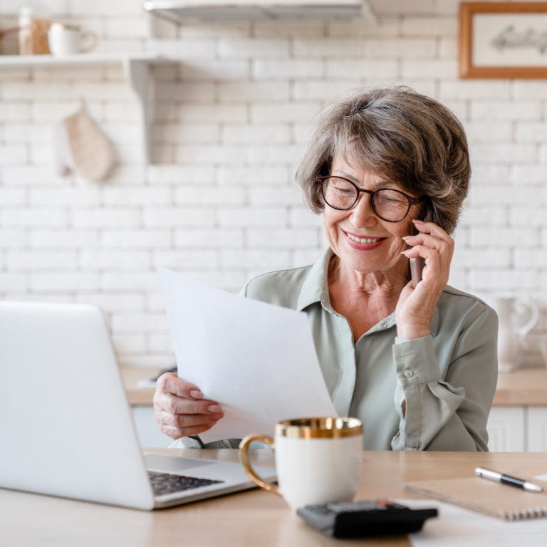 A smiling retired, elderly woman on her mobile phone, reviewing her policy details online and holding a hardcopy of her contract now managed by another insurer.