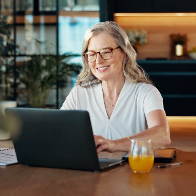 Smiling middle-aged white woman at a table with a laptop and orange juice, reviewing retirement benefits.