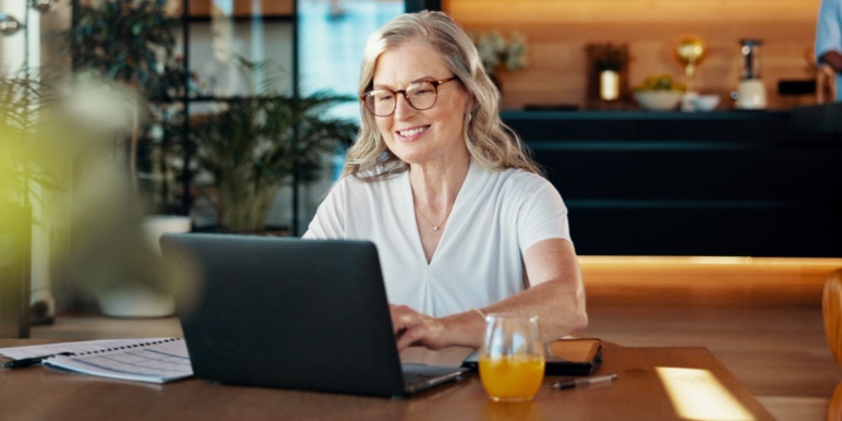 Smiling middle-aged white woman at a table with a laptop and orange juice, reviewing retirement benefits.