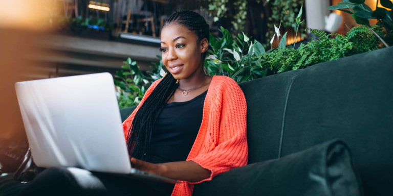 Young lady sitting on a black couch wearing a black top and an orange cardigan jersey. She's smiling while working on her laptop.