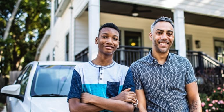 Father and son standing in front of a white car in front of their porch. 